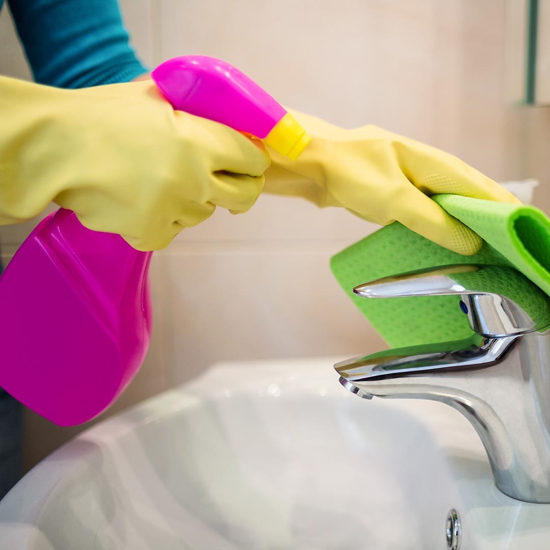 Person wearing rubber gloves and cleaning bathroom sink with a spray bottle and a sponge